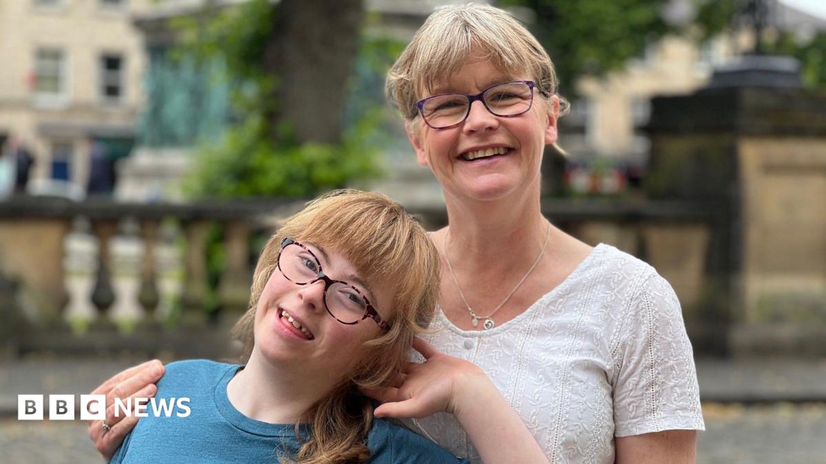 A young girl smiles with her head cocked to the side as she leans back against her mother, who is smiling and has her arm wrapped around her. The pair are stood outside.