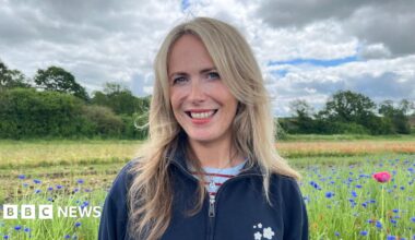 Evitts stood in a field of blue cornflowers, looking into the camera and smiling. She has long blonde hair, and is wearing a blue fleece with a white daisy logo embroidered on the chest. The sky is cloudy with some blue patches, and along the edge of the field are trees and bushes.