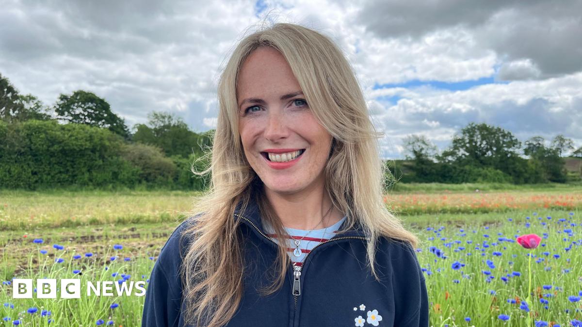 Evitts stood in a field of blue cornflowers, looking into the camera and smiling. She has long blonde hair, and is wearing a blue fleece with a white daisy logo embroidered on the chest. The sky is cloudy with some blue patches, and along the edge of the field are trees and bushes.