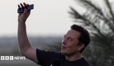 Elon Musk wearing a black T-shirt and holding his phone above his head.