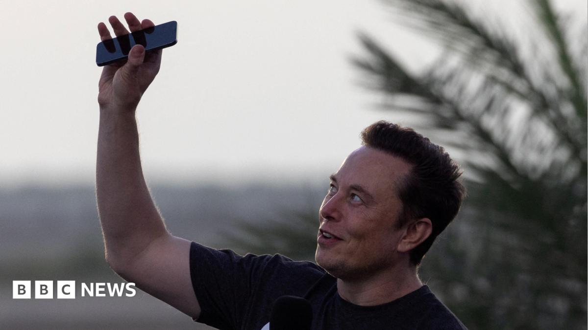 Elon Musk wearing a black T-shirt and holding his phone above his head.