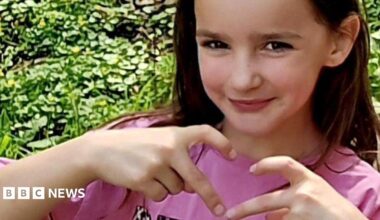 Aria Thorpe. She is pictured outside in front of a hedge. She has long dark hair and brown eyes and is looking ahead, making a heart symbol with her fingers.