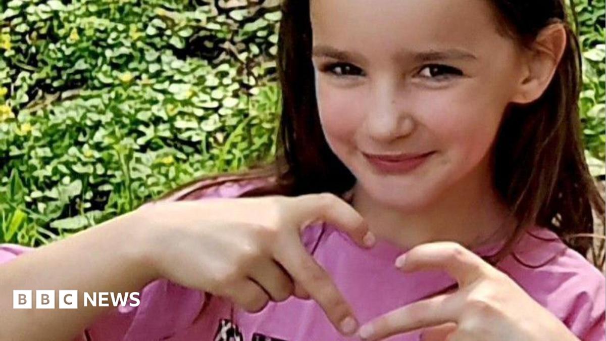 Aria Thorpe. She is pictured outside in front of a hedge. She has long dark hair and brown eyes and is looking ahead, making a heart symbol with her fingers.