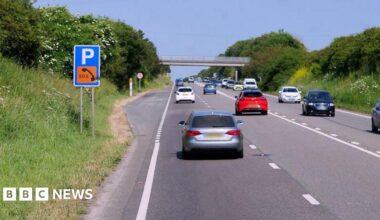 Cars travel on both sides of the A30 near Connor Downs in Cornwall on a sunny day. A layby with a sign for parking and SOS phone is on the left-hand side. A bridge crossing over the road is in the distance.