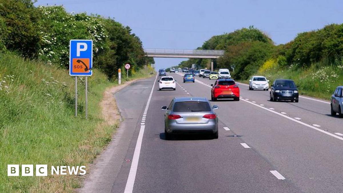 Cars travel on both sides of the A30 near Connor Downs in Cornwall on a sunny day. A layby with a sign for parking and SOS phone is on the left-hand side. A bridge crossing over the road is in the distance.