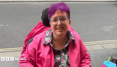 A woman with short purple hair and large blue-rimmed square glasses smiles at the camera. She sits in an electric wheelchair, wearing a shirt with a repeat skull pattern, and a bright pink jacket.