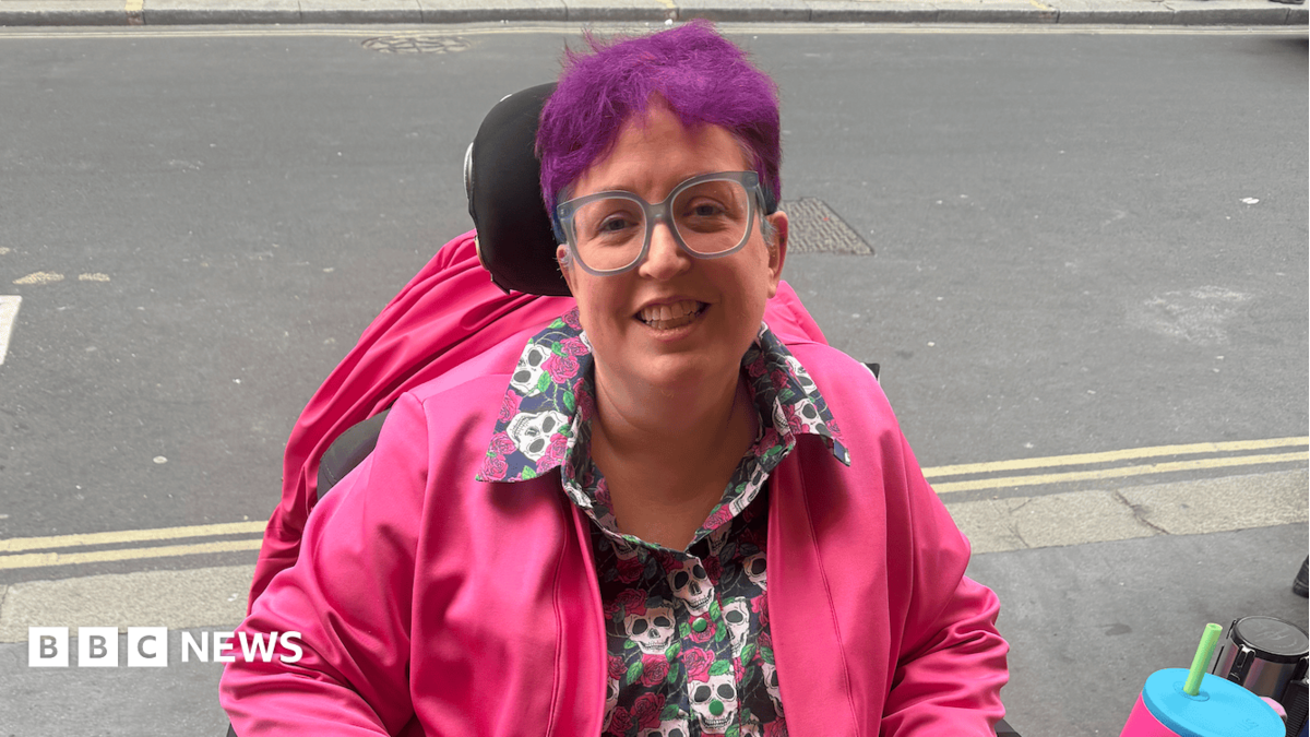 A woman with short purple hair and large blue-rimmed square glasses smiles at the camera. She sits in an electric wheelchair, wearing a shirt with a repeat skull pattern, and a bright pink jacket.