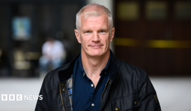 Al Carns is smiling. He has short grey hair. He's wearing an open navy shirt and a jacket. The background is blurred.