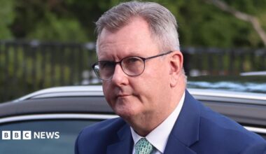 A man with short greying hair, the recededing hair line parted to the side, is getting out of a car. He is wearing black glass, a blue suit jacket, white tie and light green tie with a pattern. The top of the car behind him is black. In the background is a metal fence and some trees