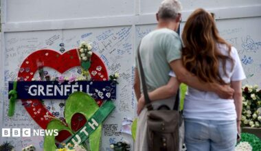 Two people hug in front of the Grenfell remembrance wall, with a heart in the background that says 'Grenfell' attached to the wall and messages people have written on the wall, in London