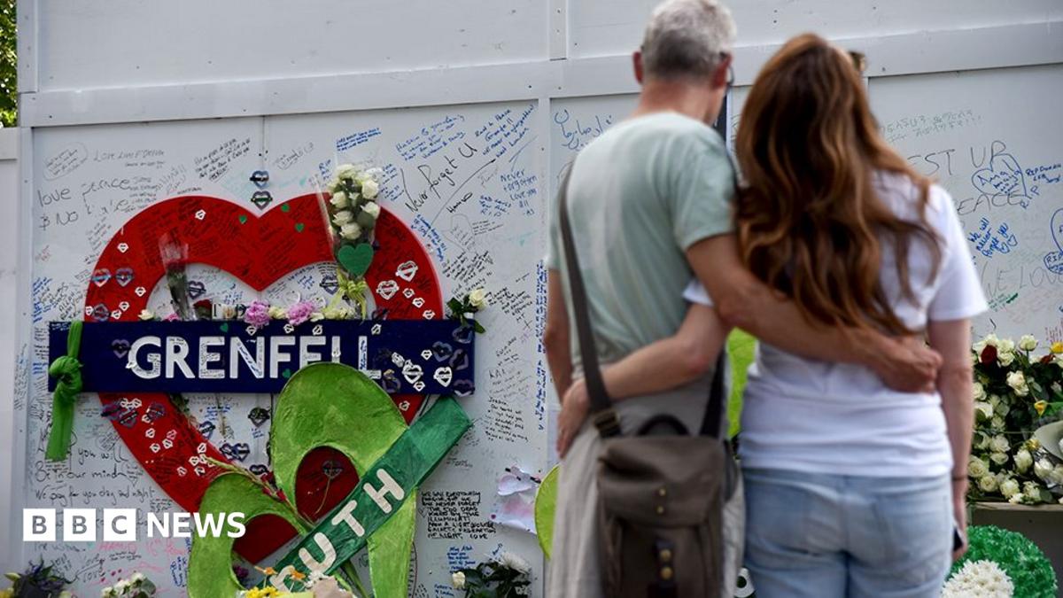 Two people hug in front of the Grenfell remembrance wall, with a heart in the background that says 'Grenfell' attached to the wall and messages people have written on the wall, in London