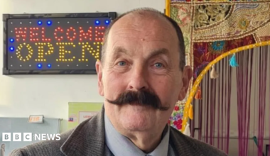 A balding man with a dark handlebar moustache is wearing a tweed jacket and standing in front of a sign with the words welcome open.