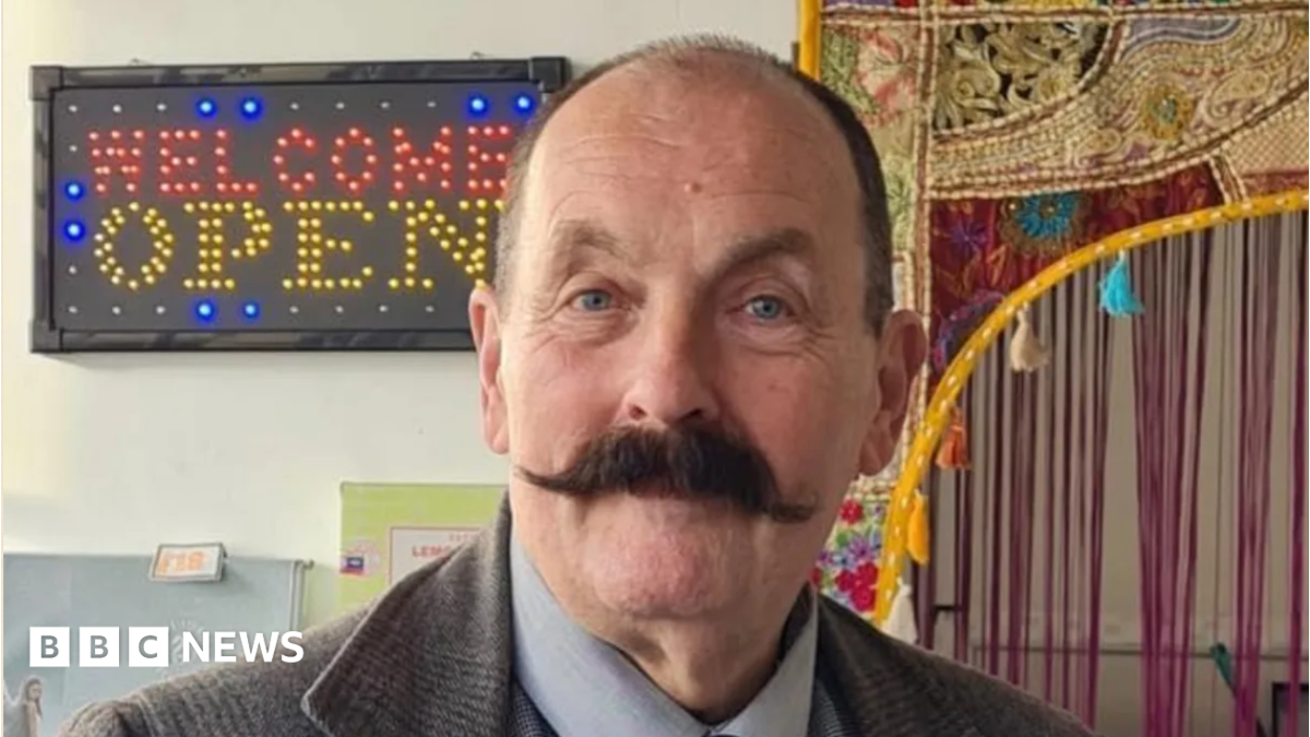 A balding man with a dark handlebar moustache is wearing a tweed jacket and standing in front of a sign with the words welcome open.