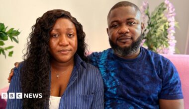 Joy Kabiri has long dark hair and is wearing a navy blue shirt with a white vest top underneath.  She is sitting next to her husband Daniel who has a beard and is wearing a navy blue t-shirt.  They are sitting on a pink sofa in their lounge.