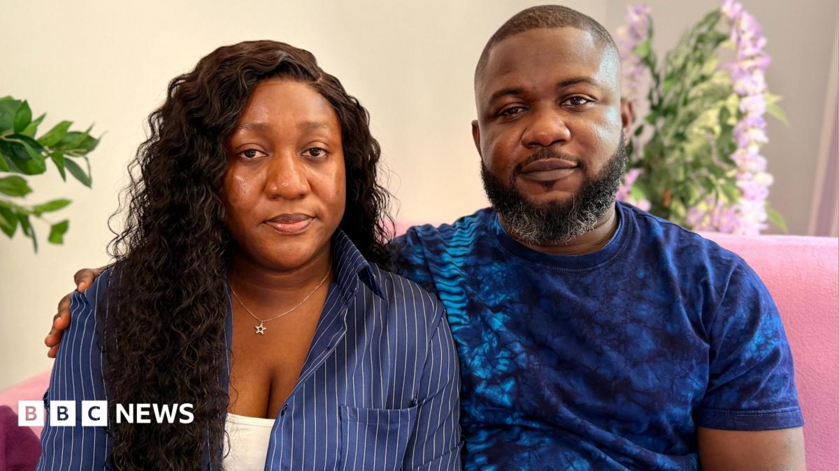 Joy Kabiri has long dark hair and is wearing a navy blue shirt with a white vest top underneath.  She is sitting next to her husband Daniel who has a beard and is wearing a navy blue t-shirt.  They are sitting on a pink sofa in their lounge.