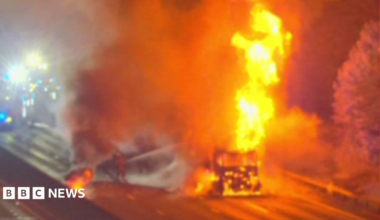 A large fire on a road, showing a lorry in flames and firefighters working to put it out. Fire engines are on the road and you can only just see their lights.
