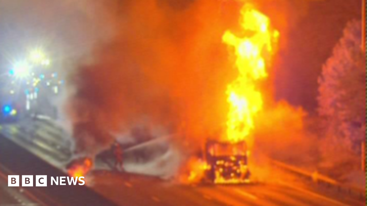 A large fire on a road, showing a lorry in flames and firefighters working to put it out. Fire engines are on the road and you can only just see their lights.