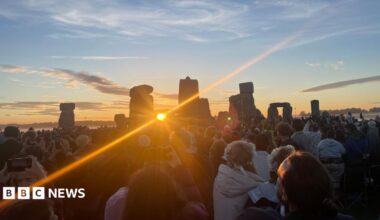 The sun is framed by two stones of Stonehenge which then reveal a strong ray of light. There are many people gathered watching the sunrise.