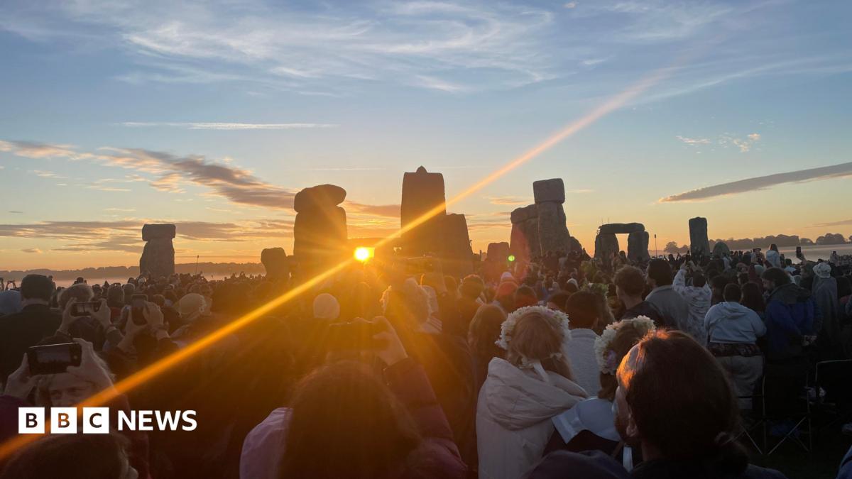 The sun is framed by two stones of Stonehenge which then reveal a strong ray of light. There are many people gathered watching the sunrise.