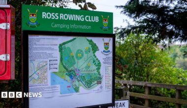 A large green information board for Ross Rowing Club stands beside a wooden footbridge, surrounded by trees and greenery.