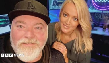 A middle-aged man with a black baseball cap and grey beard and a blonde-haired younger woman smile at the camera