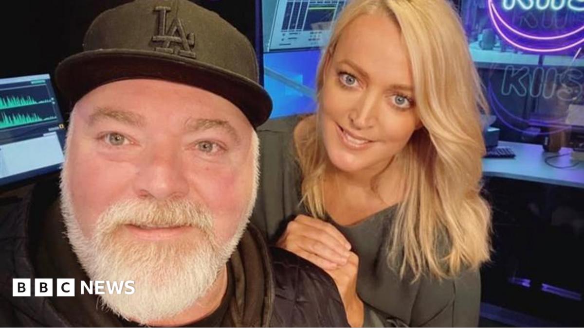 A middle-aged man with a black baseball cap and grey beard and a blonde-haired younger woman smile at the camera