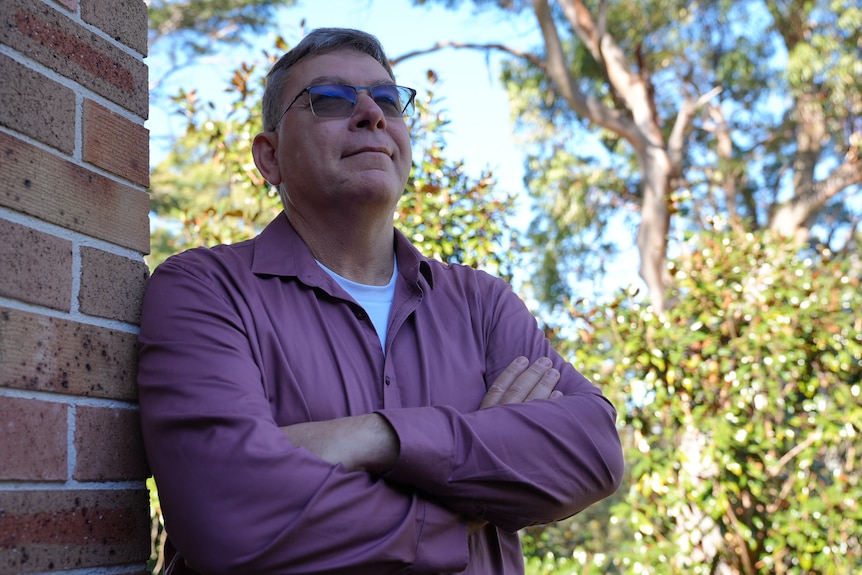 An Aboriginal man is standing next to a brick wall with his arms crossed.