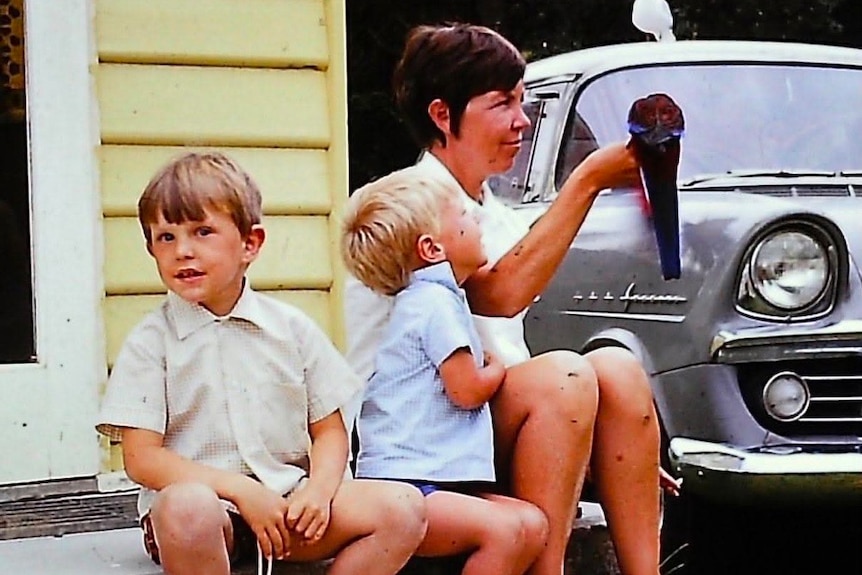 A mother sits with her two sons while they talk about the grey car nearby.