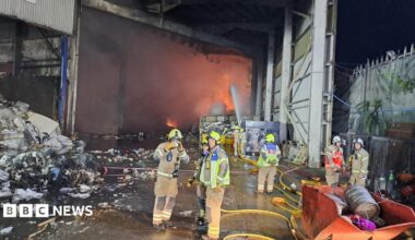 Several firefighters stand in a warehouse building, with flames visible in the background.