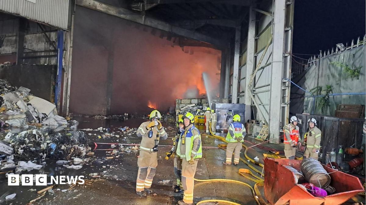 Several firefighters stand in a warehouse building, with flames visible in the background.