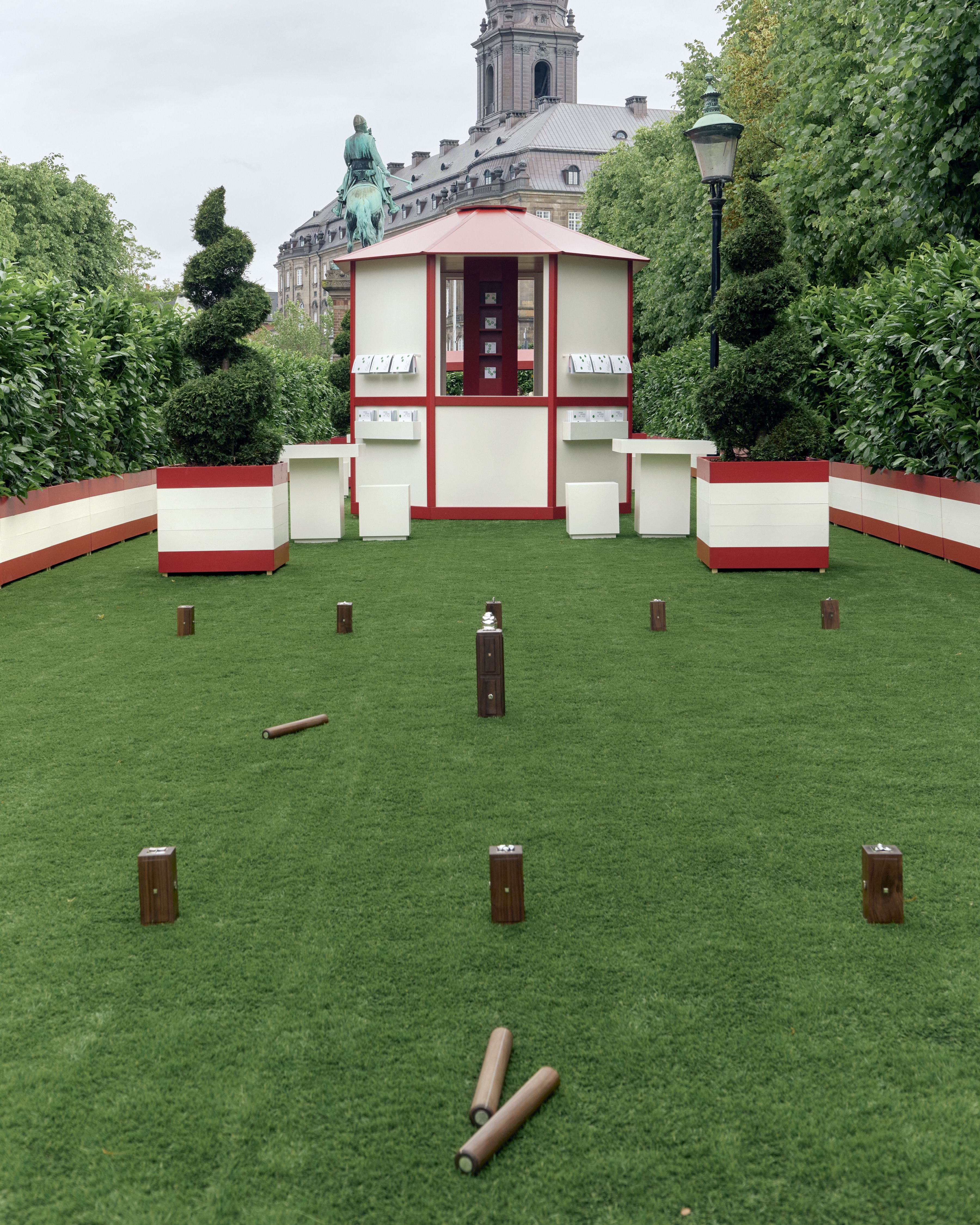 An open-air playground with green turf, wooden podiums used to display chrome games, and a wrap-around red and beige fence with matching central kiosk, all surrounded by greenery.
