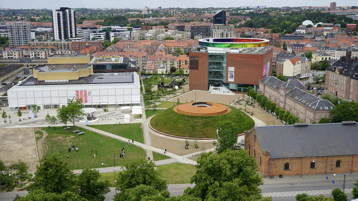 Aerial view of a city with a modern art museum, colorful rooftop rainbow, and surrounding historic buildings.