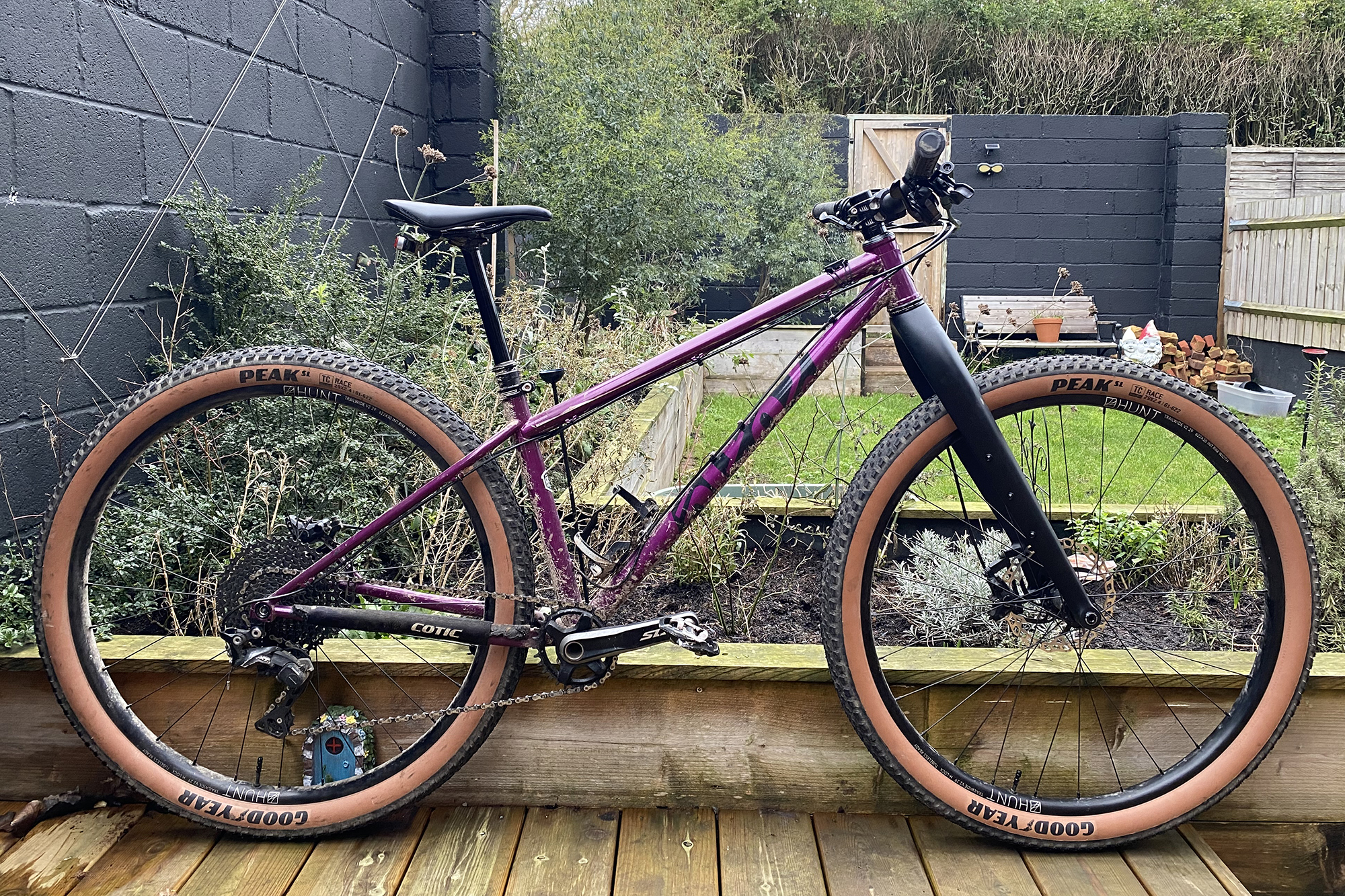 A 2018 Cotic Solaris Max in purple fitted with a rigid carbon fork, Goodyear tan wall tyres with the backdrop of a small garden