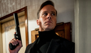 Man in a black turtleneck holds a suppressed handgun while peering cautiously around a doorway.
