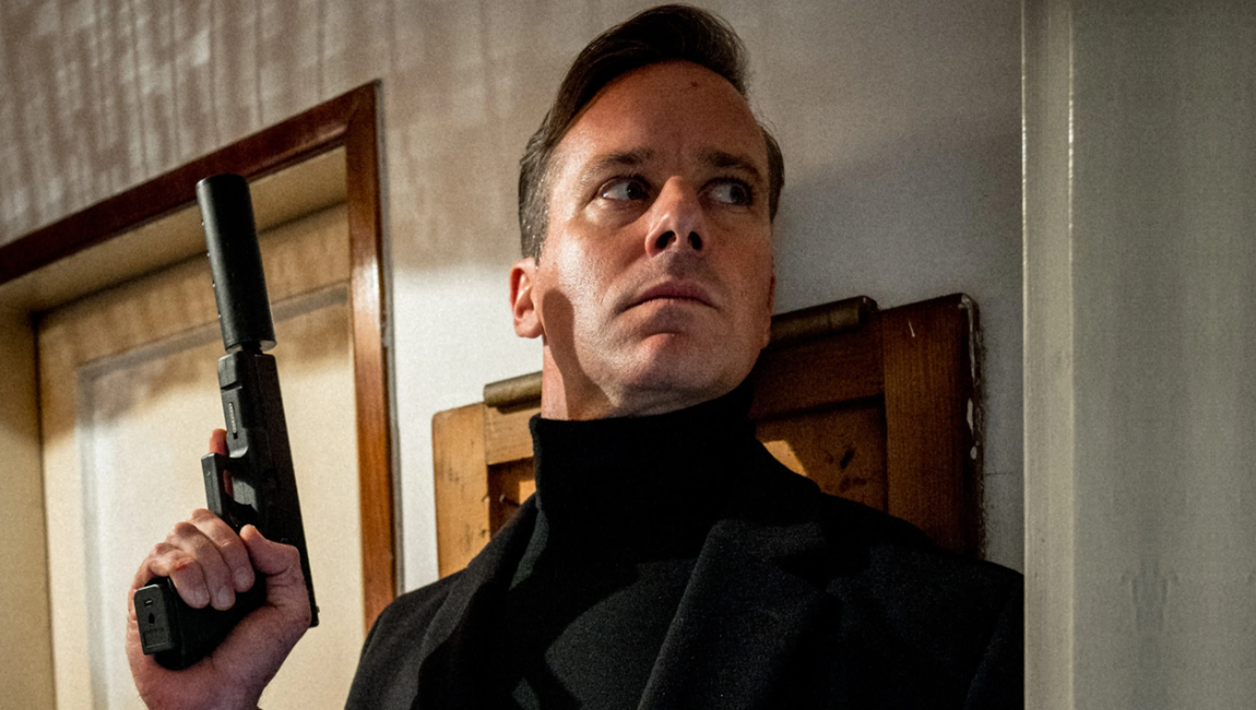Man in a black turtleneck holds a suppressed handgun while peering cautiously around a doorway.