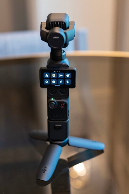 A compact handheld camera with a small display screen showing camera settings, mounted on a mini tripod, is placed on a reflective surface.