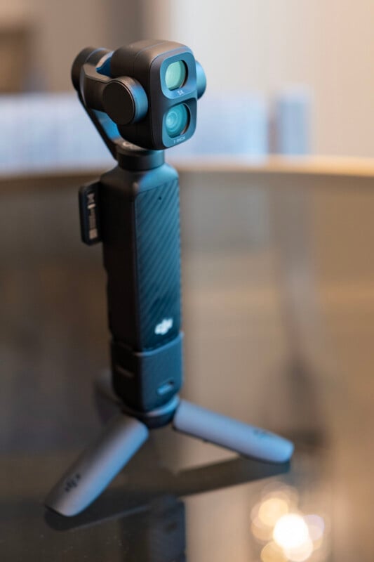 A compact DJI handheld gimbal camera with dual lenses is mounted on a small tripod and placed on a reflective glass surface indoors.