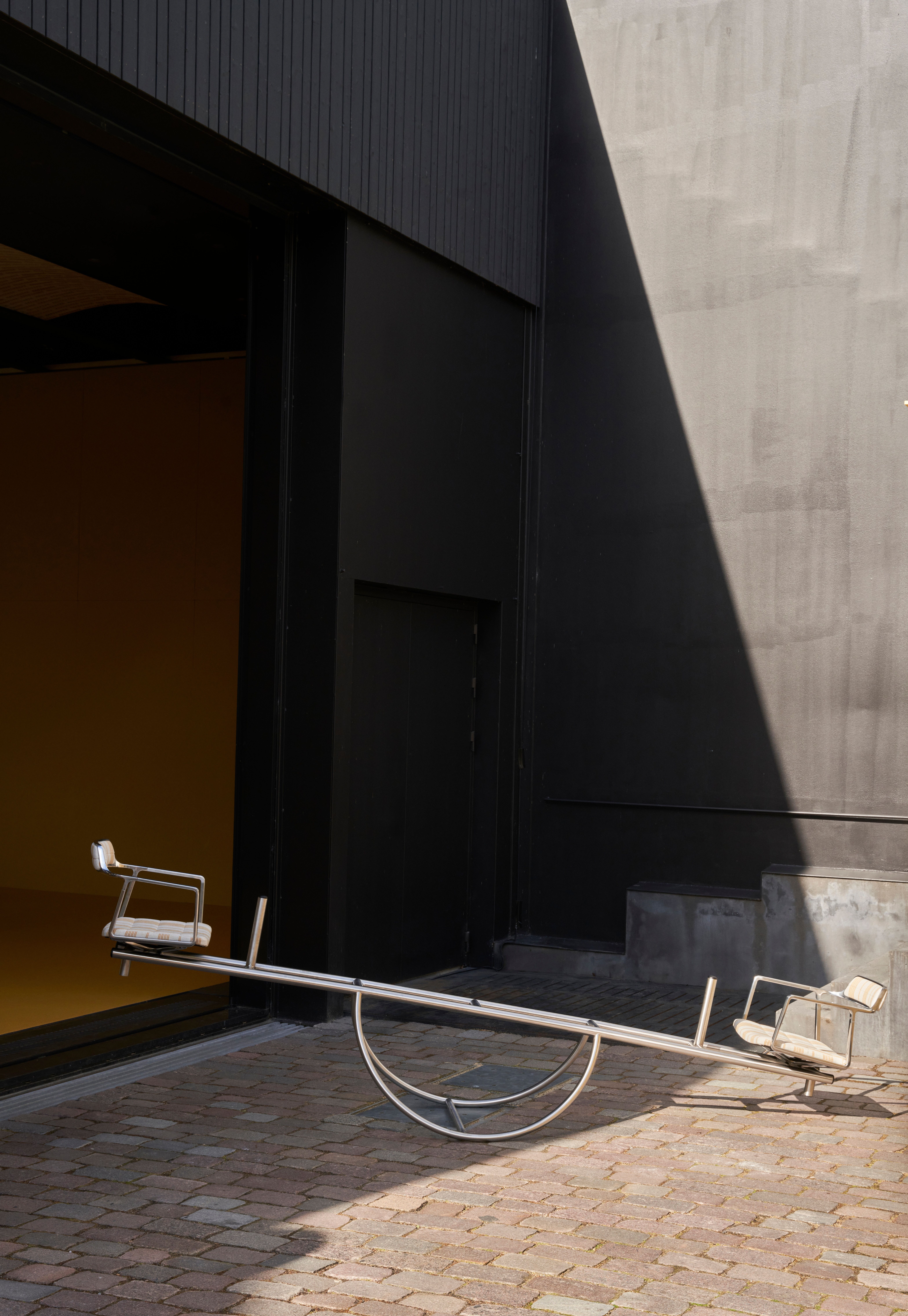 A playful installation comprising a chrome-clad, two-seat rocking chair, a butter yellow-painted kiosk, and a massive modular sofa with a square surface, butter yellow fabric, and chrome detailing.