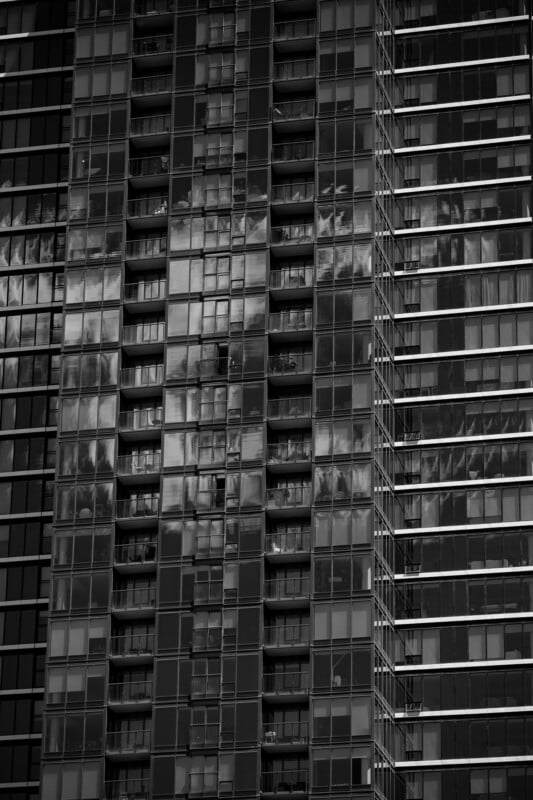 Black and white photo of a modern high-rise building facade, with glass balconies and reflective windows. Light and shadow patterns create abstract reflections across the structure.