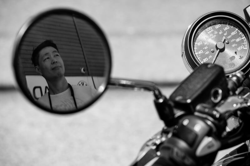 A man’s face is reflected in the round side mirror of a motorcycle, with the motorcycle's speedometer and handlebars also visible in the black-and-white image.