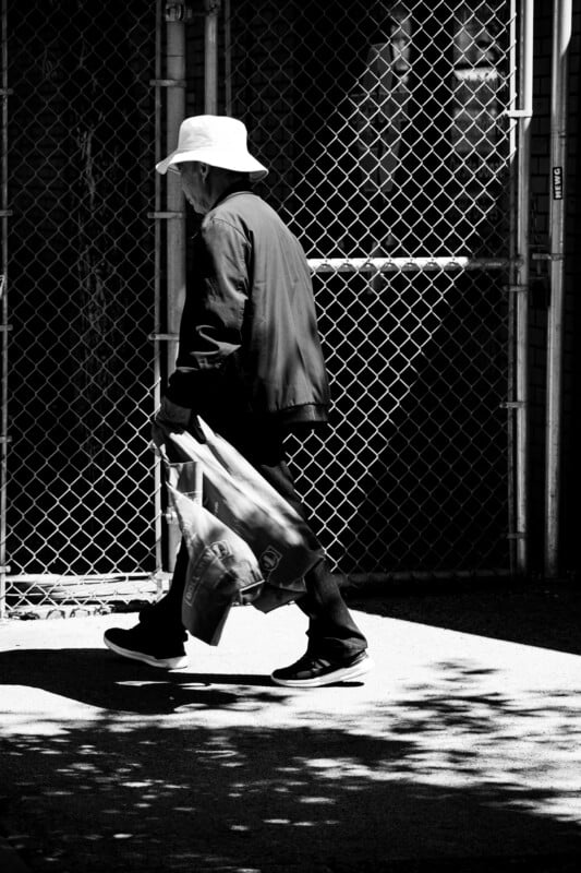 A person wearing a wide-brimmed hat and dark clothing walks along a sidewalk, carrying plastic bags in each hand. They pass by a chain-link fence, with contrasting light and shadow on the ground.