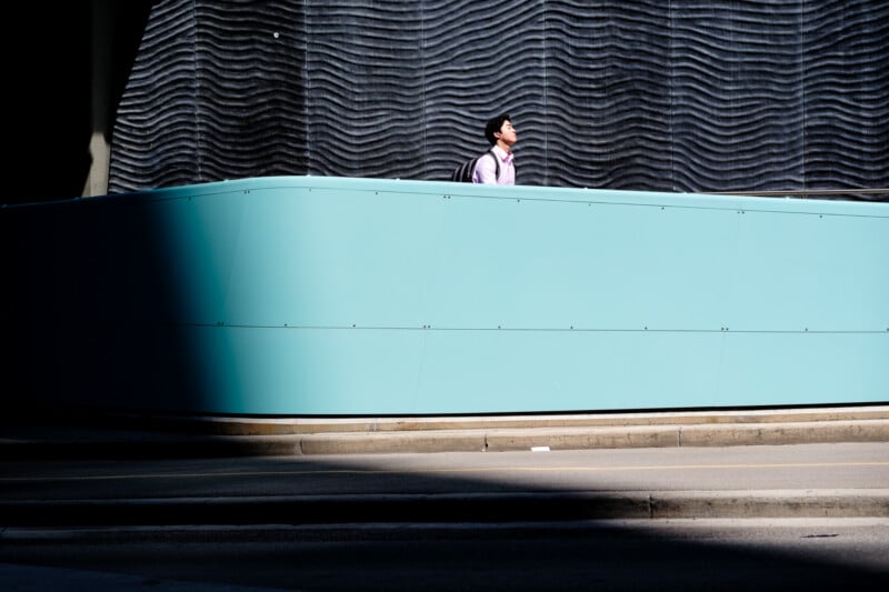 A person wearing a pink shirt and backpack walks beside a tall, turquoise wall in partial sunlight. Wavy dark grey panels are visible above the wall, creating a modern urban scene.