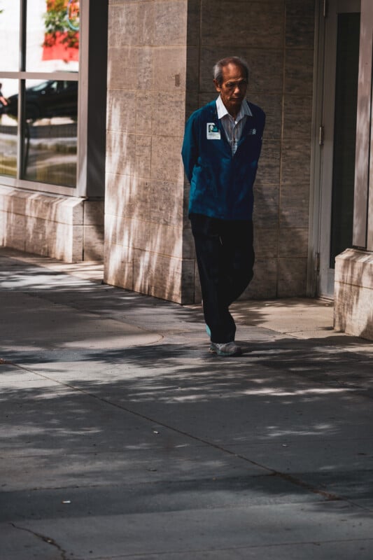 An older man in a blue jacket walks alone on a shaded city sidewalk with his hands behind his back, passing by a stone building.