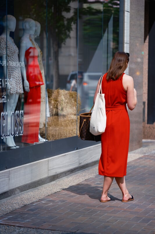 A woman in a sleeveless red dress and sandals walks on a city sidewalk, carrying a white tote bag and a brown handbag. She passes a store window displaying two mannequins in summer outfits.