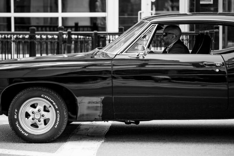 A man wearing sunglasses sits in the driver's seat of a classic black car parked on a city street. The photo is in black and white, and buildings with large windows and railings are visible in the background.
