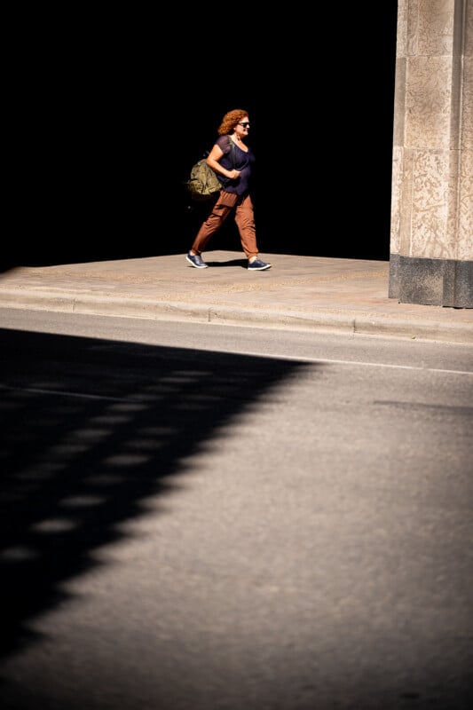 A woman with curly hair, sunglasses, and a large backpack walks on a sunlit sidewalk beside a tall stone building, with a dark shadowy background contrasting the bright street.