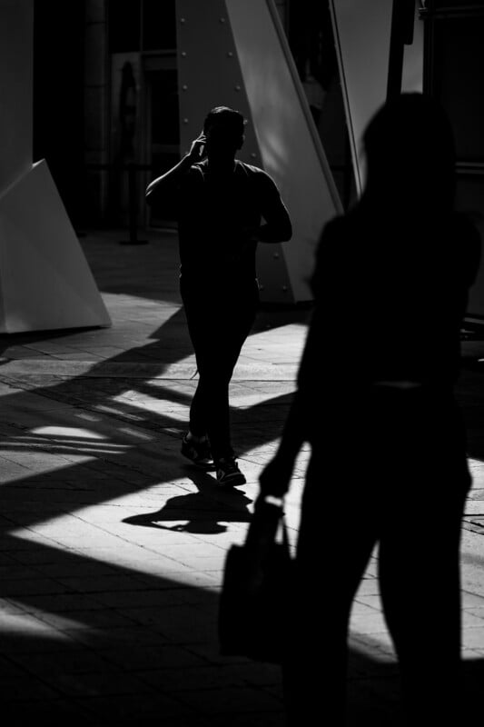 A black and white photo showing a person walking and talking on the phone in a sunlit area, while another person stands in the foreground, both partly silhouetted by the contrast between light and shadow.