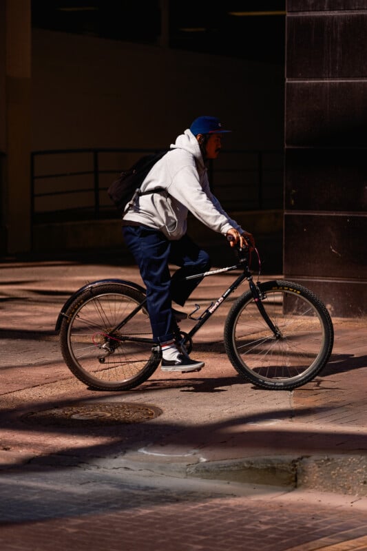 A person wearing a gray hoodie, blue pants, and a blue cap rides a black bicycle along a city street, with a backpack on their back and sunlight casting shadows on the pavement.