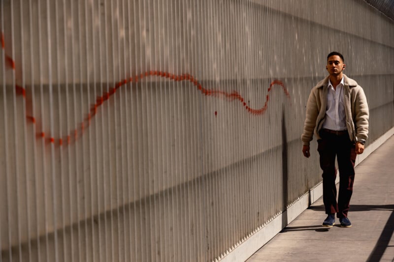A man in casual clothes walks alone along a sunlit sidewalk next to a corrugated metal wall with a red wavy line painted across it.