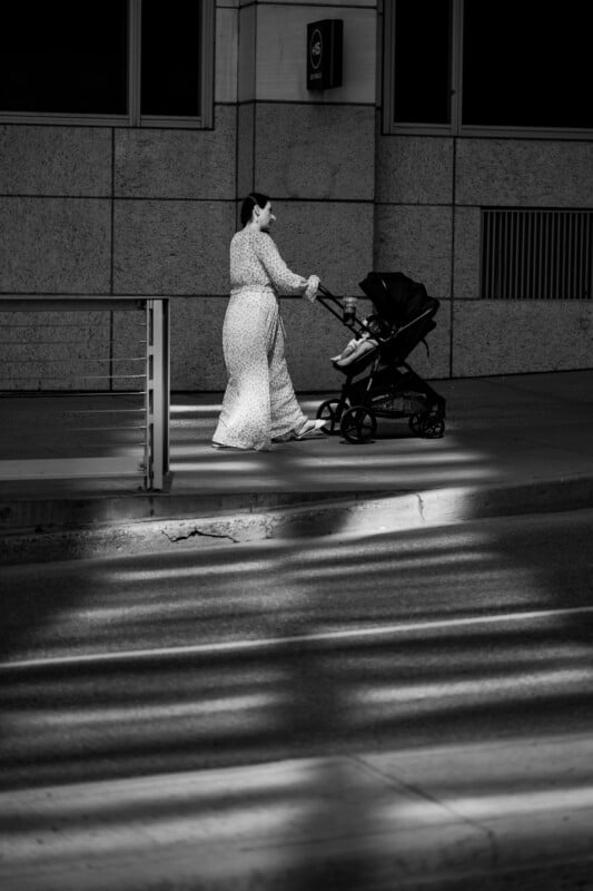 A woman in a long patterned dress pushes a stroller along a city sidewalk, crossing a sunlit patch among shadows. The background shows a modern building facade. The image is in black and white.